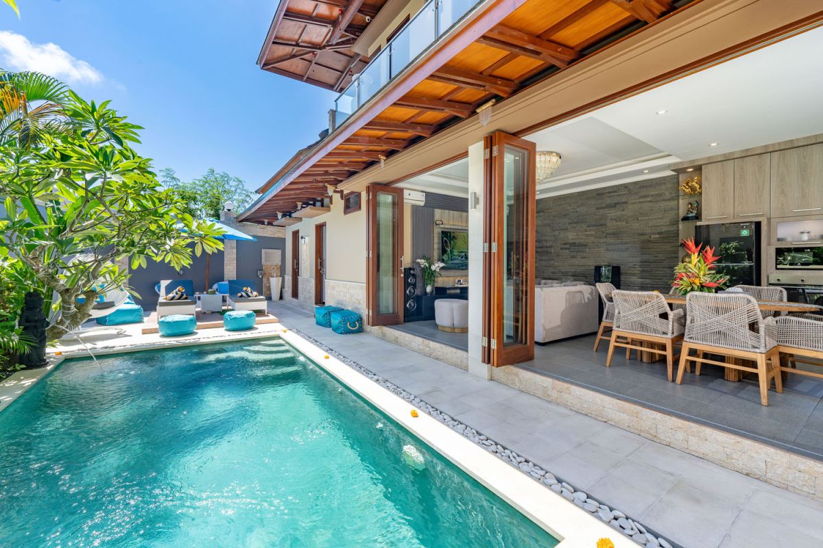 Modern Balinese villa with infinity pool, open sliding doors to lounge area, and lush frangipani trees under blue skies. - Bali Villas